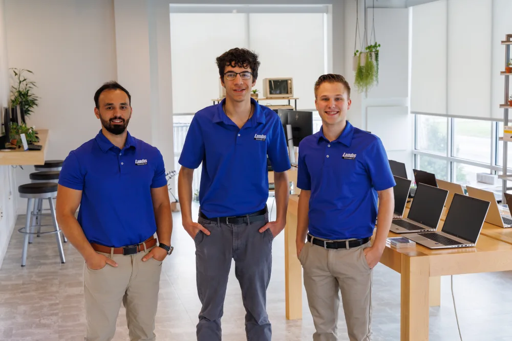 Three men in blue Landis Technologies LLC shirts standing inside an office space with laptops displayed on tables and plants in the background.