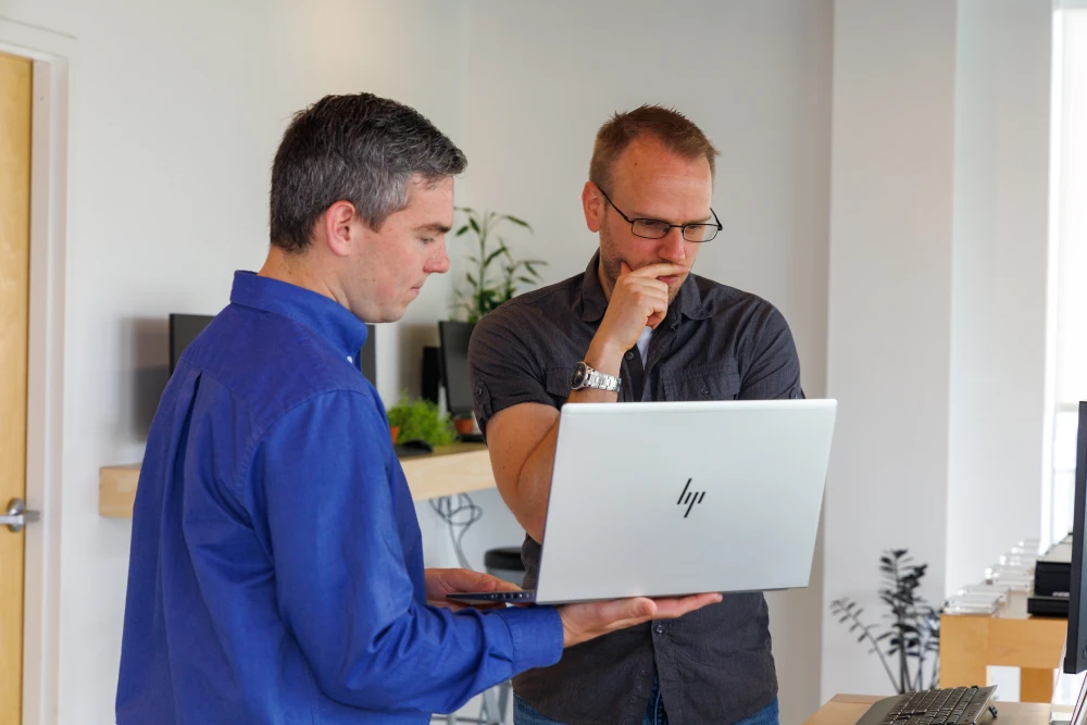 Two men in an office, one in a blue shirt holding an HP laptop, the other in a black shirt looking at the screen thoughtfully.