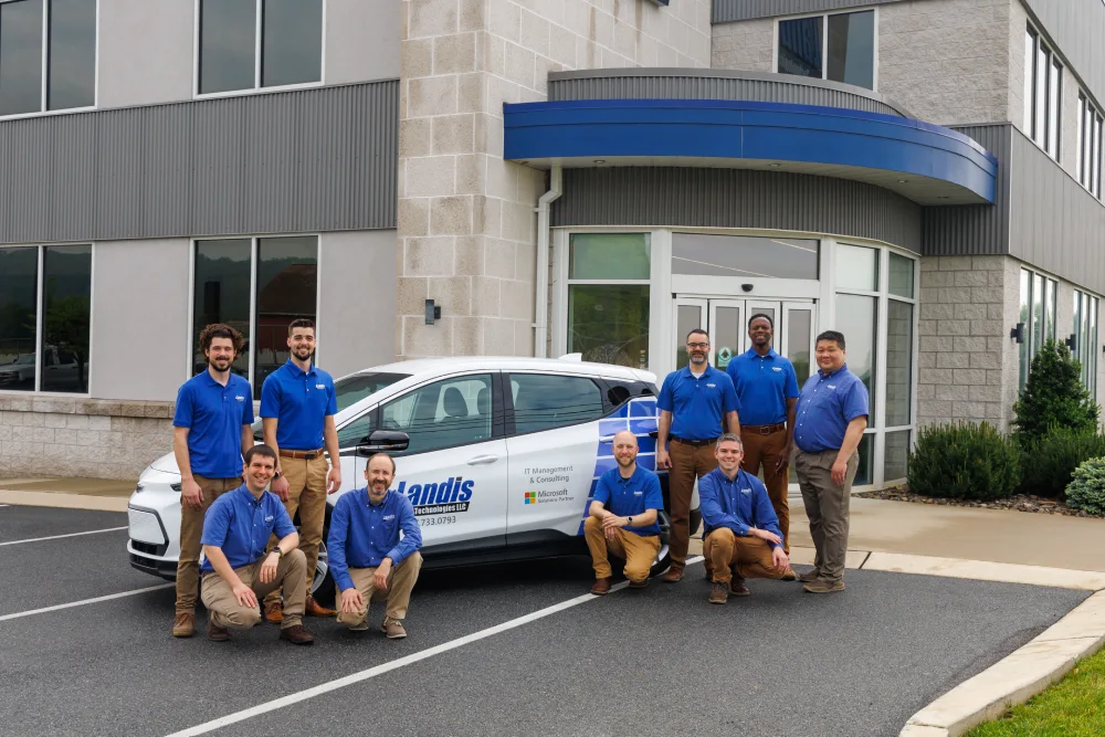 A group of nine men in blue shirts and khaki pants standing and kneeling in front of a Landis Technologies LLC vehicle parked outside Landis office building.