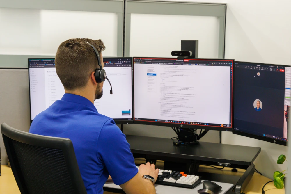 Man in a blue shirt and headset working at a desk with three monitors displaying various applications in an office cubicle.